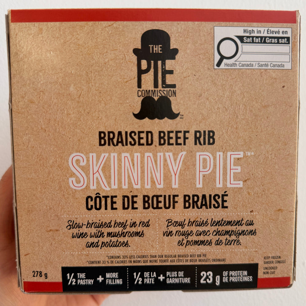 A hand holding a 223 g box of The Pie Company's Skinny Pie Braised Beef Rib, shown against a plain white wall.