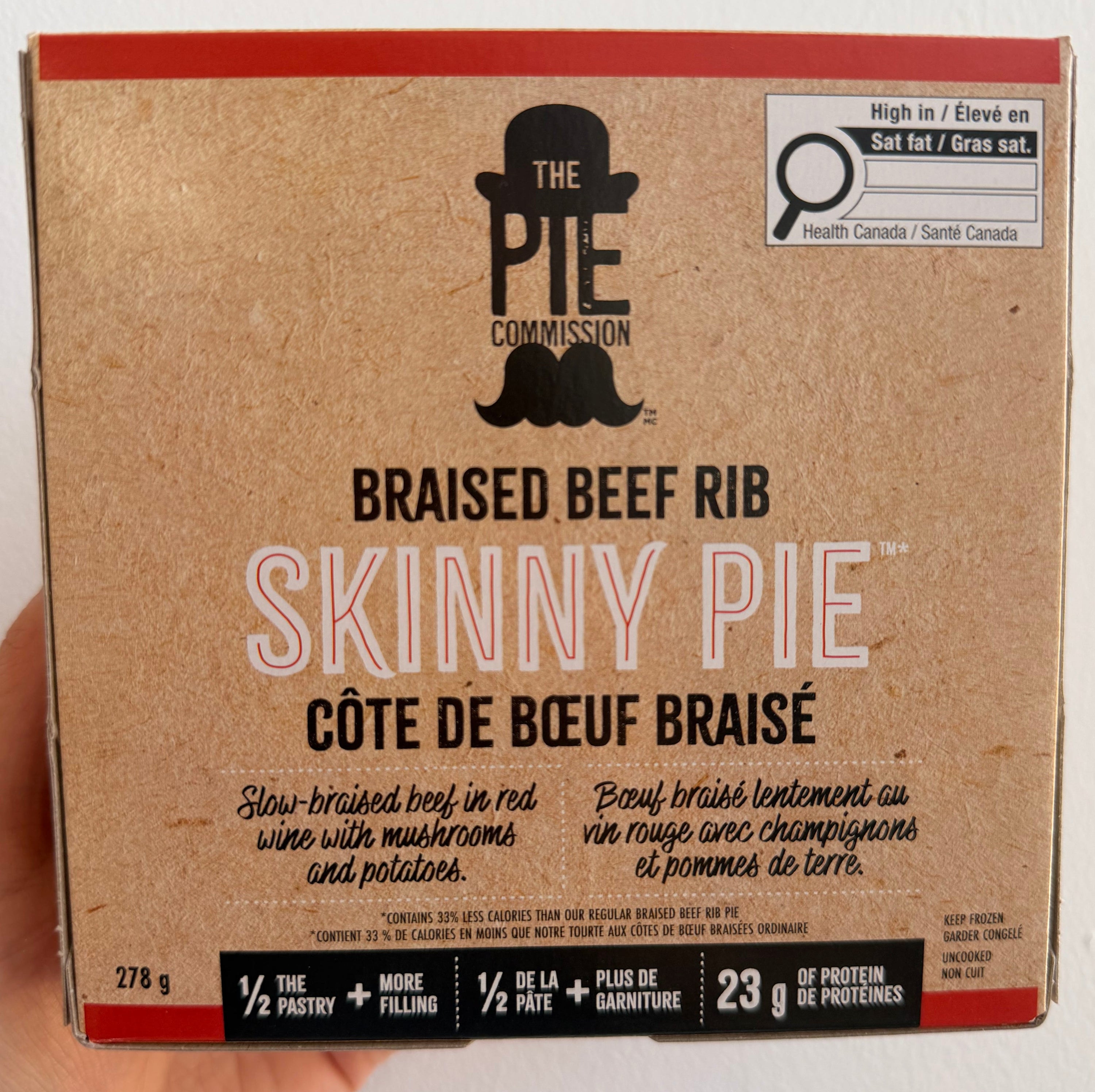 A hand holding a 223 g box of The Pie Company's Skinny Pie Braised Beef Rib, shown against a plain white wall.